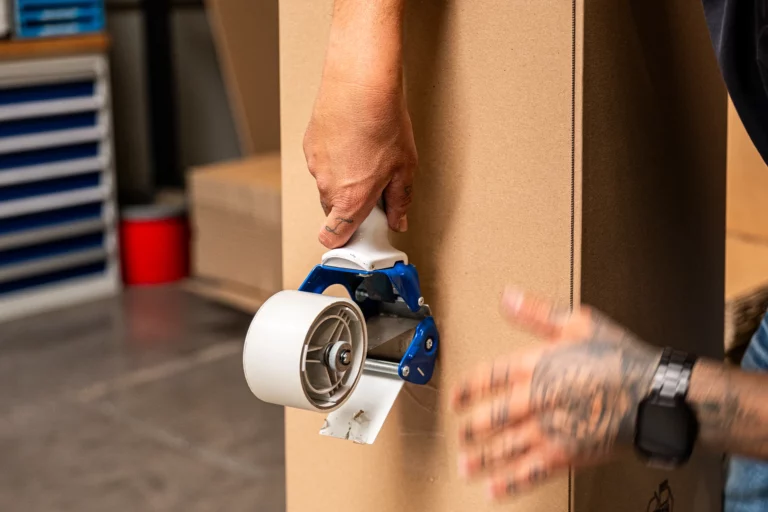 Eine Hand benutzt einen Paketbandabroller, um einen Karton zu verschließen.